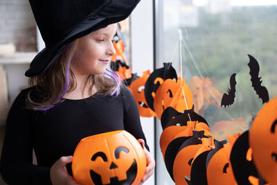 Little girl in costume of witch holding pumpkin jack with candies, celebrating halloween at home