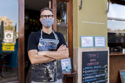 Male owner in face mask standing with arms crossed against cafe during covid-19 crisis