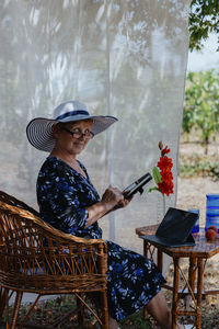 Elderly woman talking on smartphone while working with tablet in garden office.