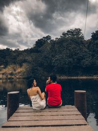 Rear view of couple sitting on pier at lake against sky