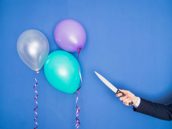 Cropped hand of man holding knife by colorful balloons against blue background