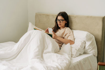 Female pretty student reading a book and taking notes on mobile phone while lying in bed