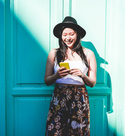 Smiling young woman using smart phone while standing against wall