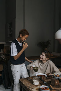 Smiling non-binary person brushing teeth by friend having breakfast at home