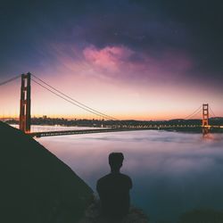 Rear view of man sitting against golden gate bridge during sunset