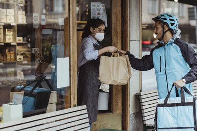 Young delivery man collecting order from female owner at delicatessen shop during covid-19