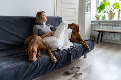Depressed sad woman sitting with two dog at home on sofa drinking tea, thinking, has mental problems