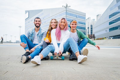 Group of teenagers fooling about at skate park - happy young friends sitting on skateboard 