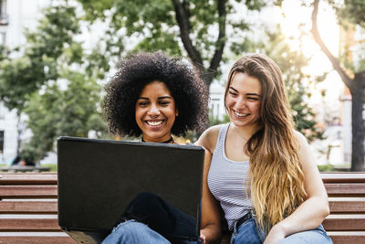 Happy friends using laptop while sitting bench