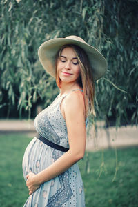 Smiling pregnant woman touching abdomen while standing on grass