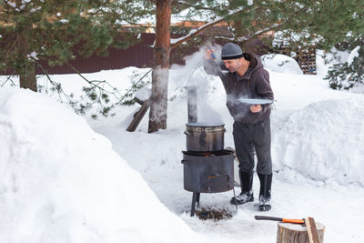 A man prepares food on a fire in winter outdoors near snowdrifts and pines, tastes soup from a ladle