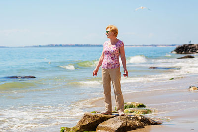 Senior woman in sunglasses stands on the beach and looks at the sea