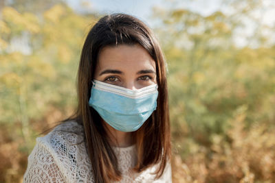 Calm female wearing protective medical mask standing in field on sunny day and looking at camera during coronavirus pandemic