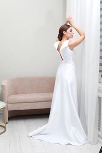 Full length portrait of bride in elegant wedding dress posing with arms raised, pensive expression 