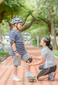 Mother assisting son in wearing kneepad on footpath