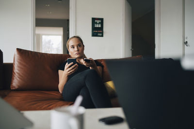 Teenage girl sitting on sofa using smart phone and remote control while watching tv at home