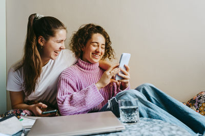 Cheerful young female friends sharing smart phone while sitting at home