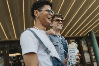 Low angle view of happy young man in sunglasses leaning on male friend holding wrap sandwich outside shop