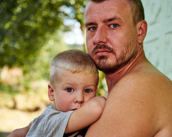 Portrait of shirtless father with son against tree