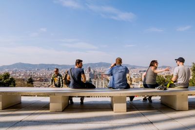 Friends sitting on railing by cityscape against sky