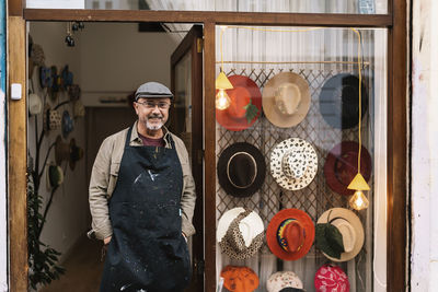 Content mature bearded male master in apron standing near showcase with collection of stylish handmade hats and looking at camera