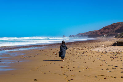 Rear view full length of woman walking at beach against clear blue sky