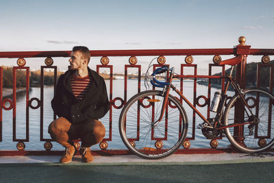 Man looking away while crouching by bicycle on bridge over river against sky in city