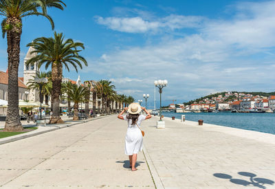 Rear view of woman wearing white sundress, walking on waterfront promenade in trogir, croatia