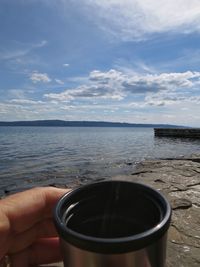 Close-up of hand holding drink by sea against sky