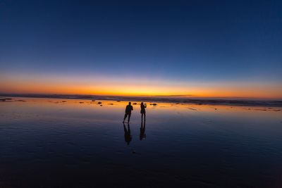 Silhouette people standing on shore at beach against dramatic sky during sunset