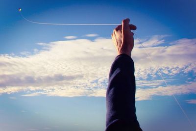 Cropped hand flying kite against cloudy blue sky on sunny day