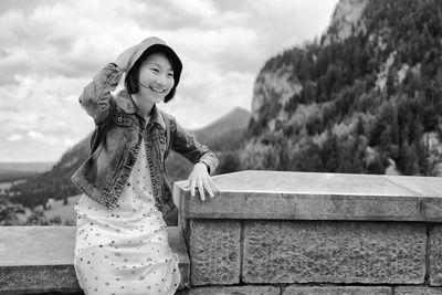 Close-up of smiling young woman sitting on retaining wall against sky