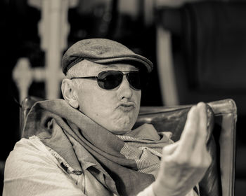 Close-up of senior man in sunglasses and cap gesturing while sitting on chair