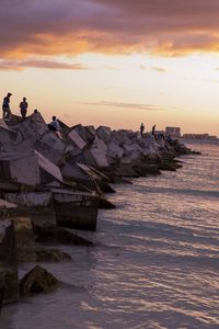 People on rocks by sea against sky during sunset