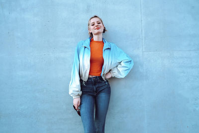 Smiling young woman with hand on hip standing in front of blue wall