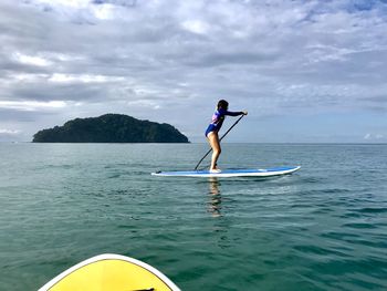 Full length of girl paddleboarding in sea against cloudy sky