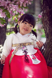 Korean girl child in a national costume sit on tree branch in a garden with cherry blossoms