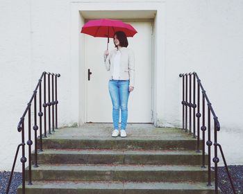 Full length of young woman holding umbrella while standing on steps against closed door