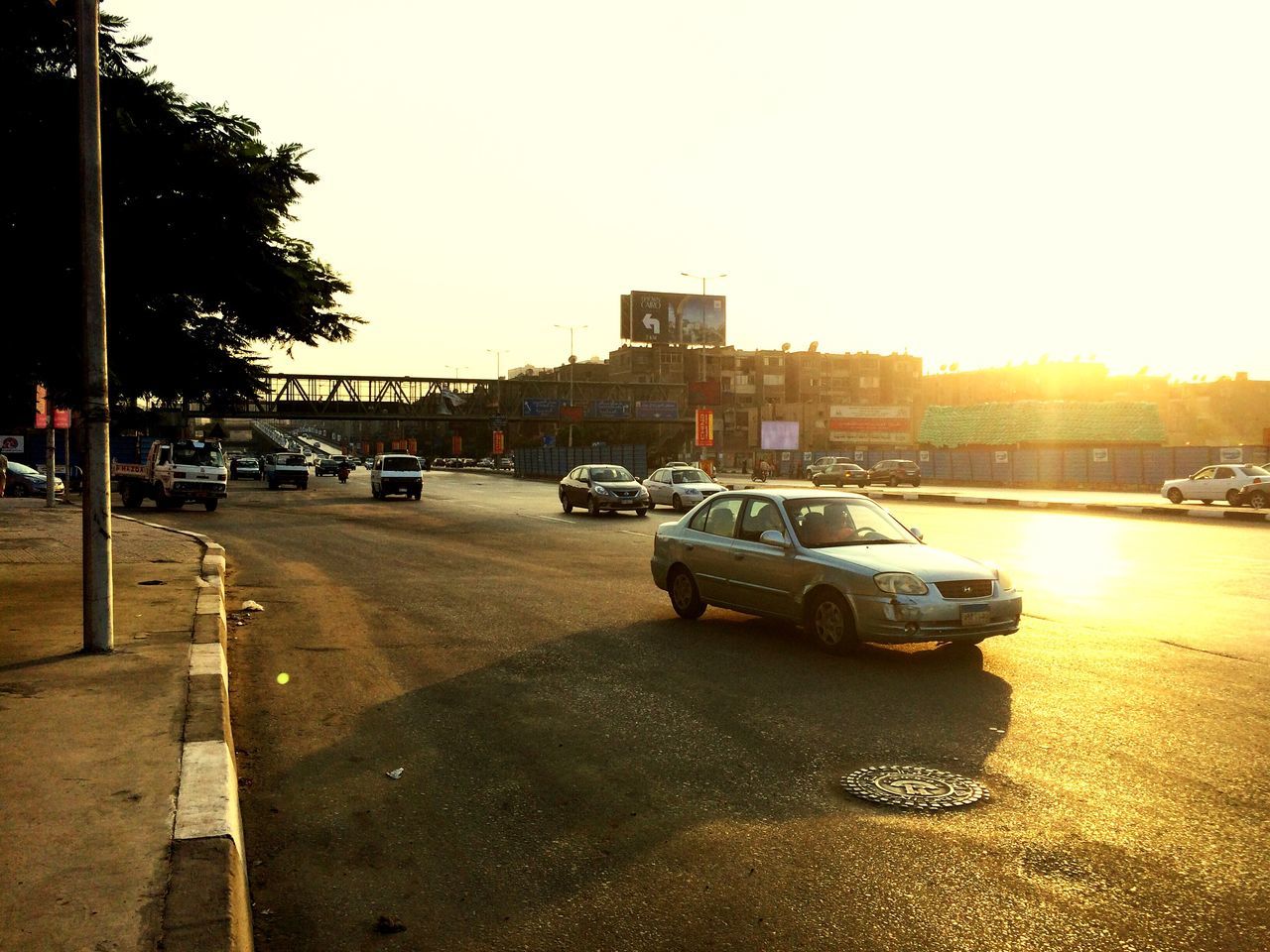 car, transportation, land vehicle, architecture, built structure, mode of transport, building exterior, street, city, road, traffic, sunbeam, sun, clear sky, sunlight, lens flare, city life, city street, outdoors, bright, development, distant