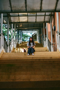 Portrait of young woman sitting on staircase while holding sunglasses at building