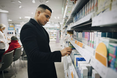 Side view of male customer choosing medicine from rack at pharmacy store