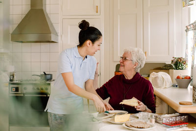 Smiling retired elderly woman talking with female caregiver in kitchen at nursing home