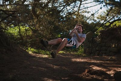 Full length of smiling young man swinging from rope over field at forest