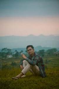 Young man sitting on field against mountains during sunset