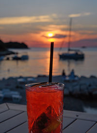 Red cocktail, focus on foreground, sunset at sea in background.