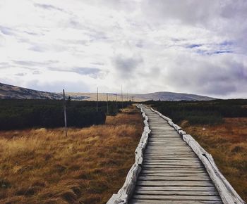 Footpath by grassy field against cloudy sky