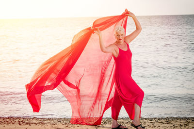 Portrait of smiling woman holding red fabric at beach during sunset