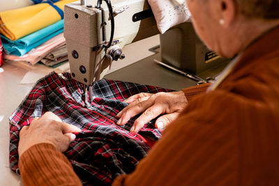 Cropped unrecognizable senior seamstress sitting at sewing machine and stitching piece of cloth while working in workshop