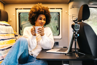 Cheerful young african american female traveler drinking coffee and enjoying video chat via laptop while resting inside camper van during summer adventure