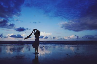 Silhouette woman standing while holding textile at beach against sky during sunset